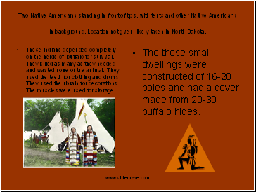 Two Native Americans standing in front of tipis, with tents and other Native Americans in background. Location not given, likely taken in North Dakota.
